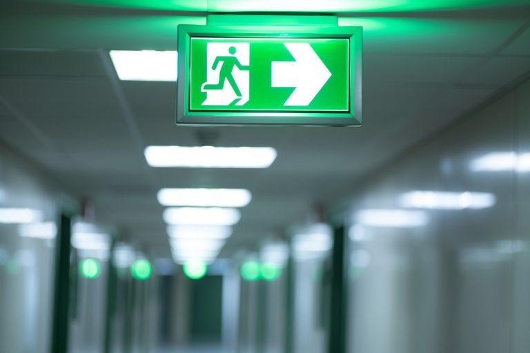 Ein grünes, beleuchtetes Notausgangsschild in einem langen, hell beleuchteten Flur. Das Schild zeigt einen laufenden Mann und einen Pfeil nach rechts, der den Fluchtweg im Notfall anzeigt.